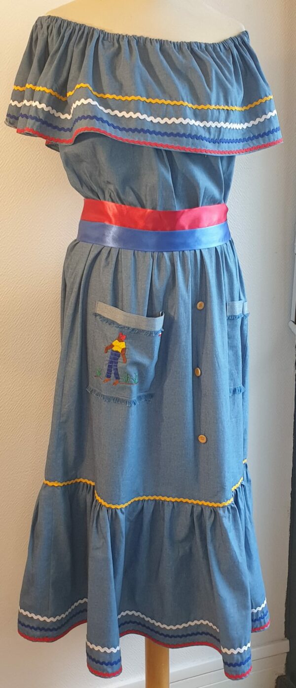 Robe Carabela cintrée sur un fond bleu et sur une tenue traditionnelle haïtienne. Deux poches sur le devant de la robe brodées à la main avec le monsieur sur la poche de droite et un petit habitat ancestral sur celle de gauche. Cette robe est conçu pour être élastique à la taille. Elle est assortie de trois boutons bois coco alignés verticalement sur le devant. Elle est aussi garnie de serpentines aux couleurs vives et elle est livrée avec une ceinture bicolore au drapeau haïtien. Créateur Anjero.