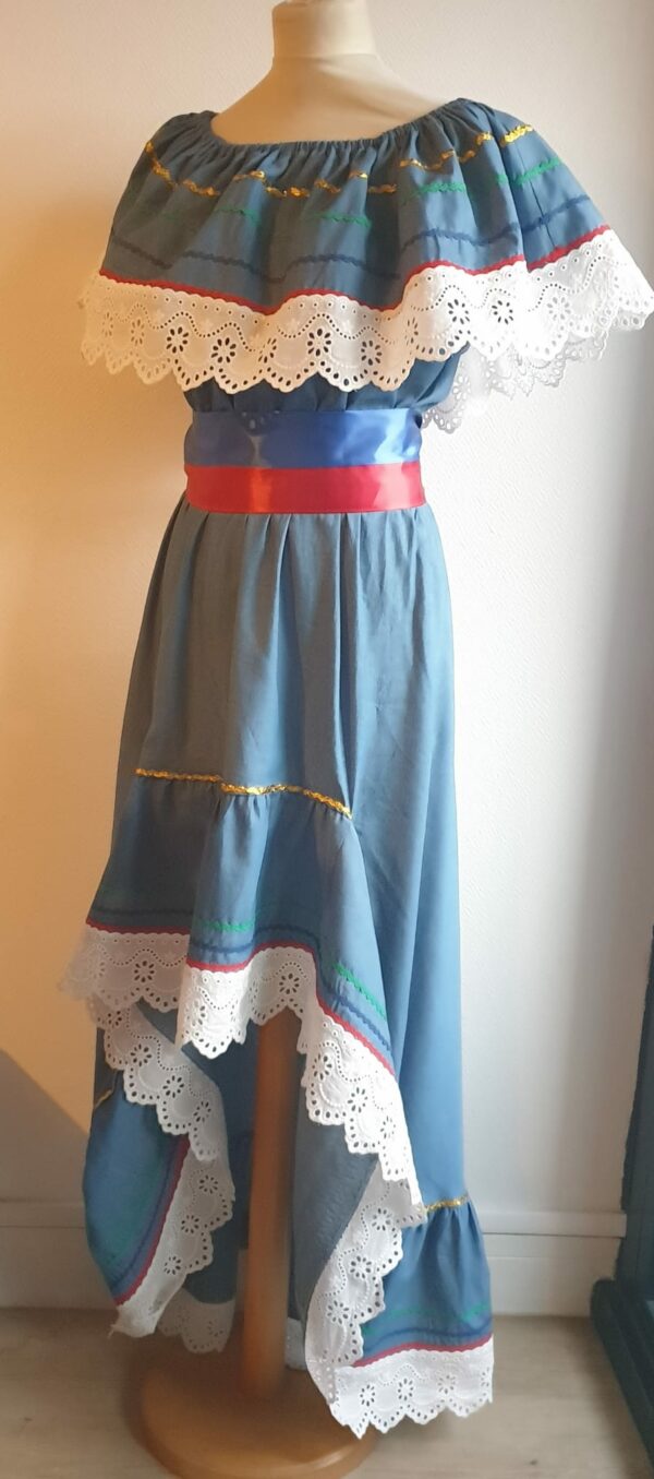 18.1 Robe Carabela cintrée sur fond bleu et sur une tenue traditionnelle haïtienne pour cérémonie, mariage, gala, baptême. Une finition au col de la robe et sur son bas de jupe est brodé en dentelles anglaises. Des serpentines aux quintuples couleurs or, vert, jaune, rouge et blanc; Une ceinture aux couleurs du drapeau haïtien; Un tissu cent pour cent coton; Une souplesse à la taille à laquelle est intégré un élastique sont autant de particularités qui confèrent à la confection toute son originalité. Made in Paris. Créé par Anjero.