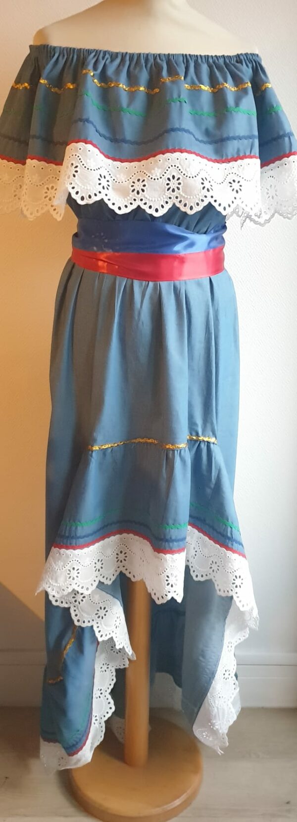 Robe Carabela cintrée sur fond bleu et sur une tenue traditionnelle haïtienne pour cérémonie, mariage, gala, baptême. Une finition au col de la robe et sur son bas de jupe est brodé en dentelles anglaises. Des serpentines aux quintuples couleurs or, vert, jaune, rouge et blanc; Une ceinture aux couleurs du drapeau haïtien; Un tissu cent pour cent coton; Une souplesse à la taille à laquelle est intégré un élastique sont autant de particularités qui confèrent à la confection toute son originalité. Made in Paris. Créé par Anjero.