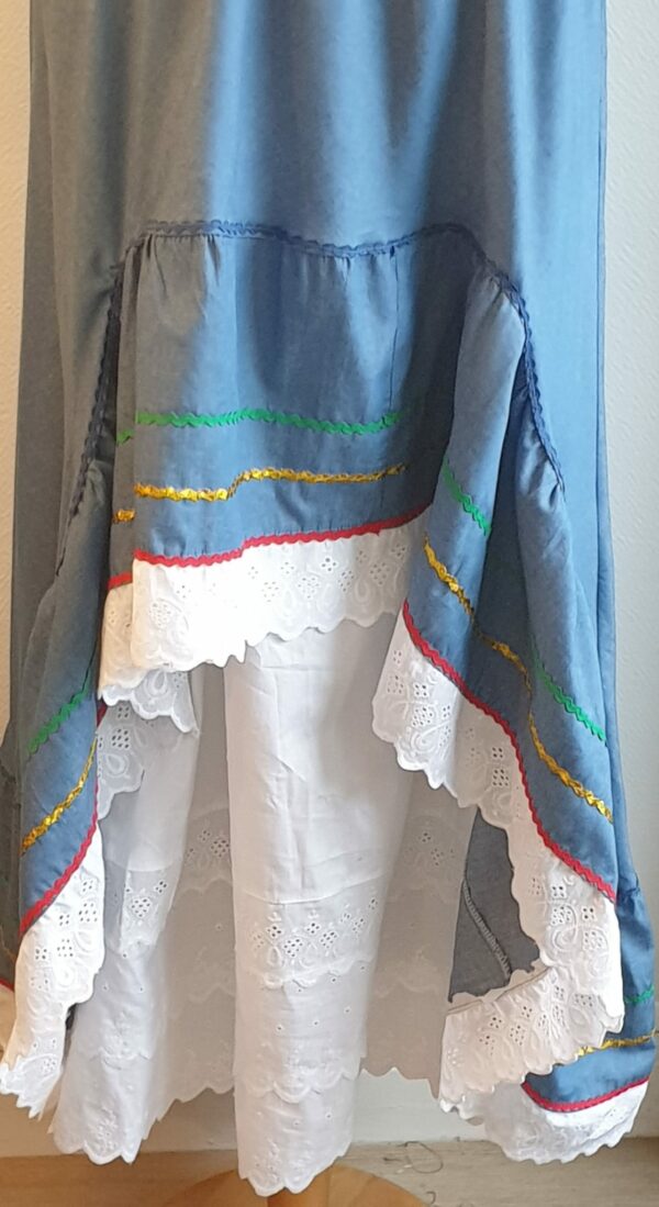 Robe Carabela cintrée sur fond bleu et sur une tenue traditionnelle haïtienne pour cérémonie, mariage, gala, baptême. Une finition au col de la robe et sur son bas de jupe est brodé en dentelles anglaises. Un jupon blanc garni de dentelles soigneusement confectionnées dans la robe, des serpentines aux quadruples couleurs jaune, bleu, blanc, rouge; Une grande ceinture aux couleurs du drapeau haïtien; Un tissu cent pour cent coton; Une souplesse à la taille à laquelle est intégré un élastique sont autant de particularités qui confèrent à la confection une entière originalité. Made in Paris. Créé par Anjero.